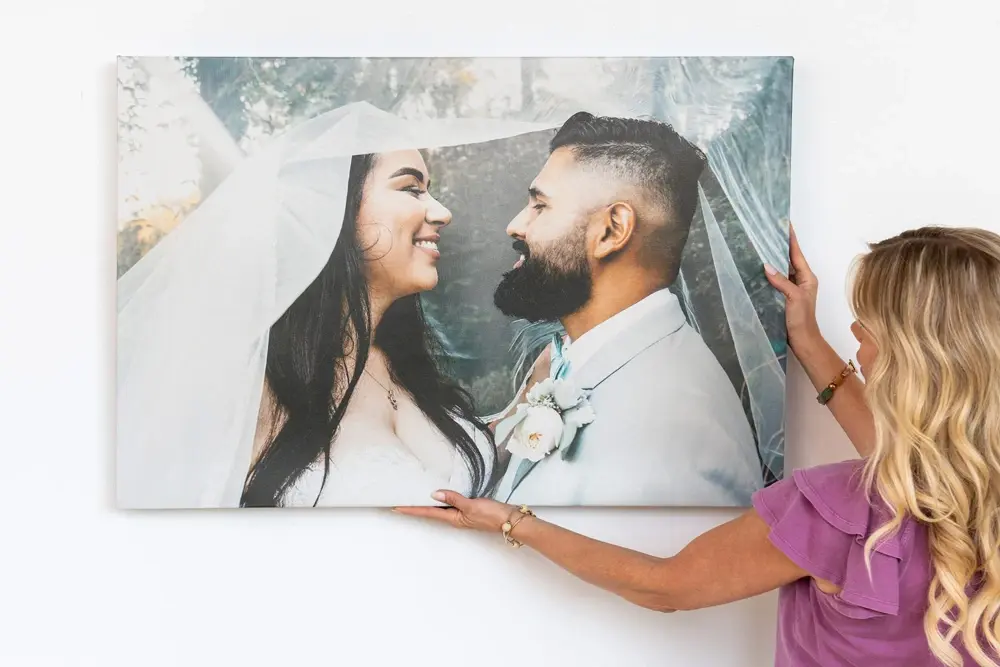 A woman in a purple top hangs a large canvas print of a smiling bride and groom under a veil on a white wall
